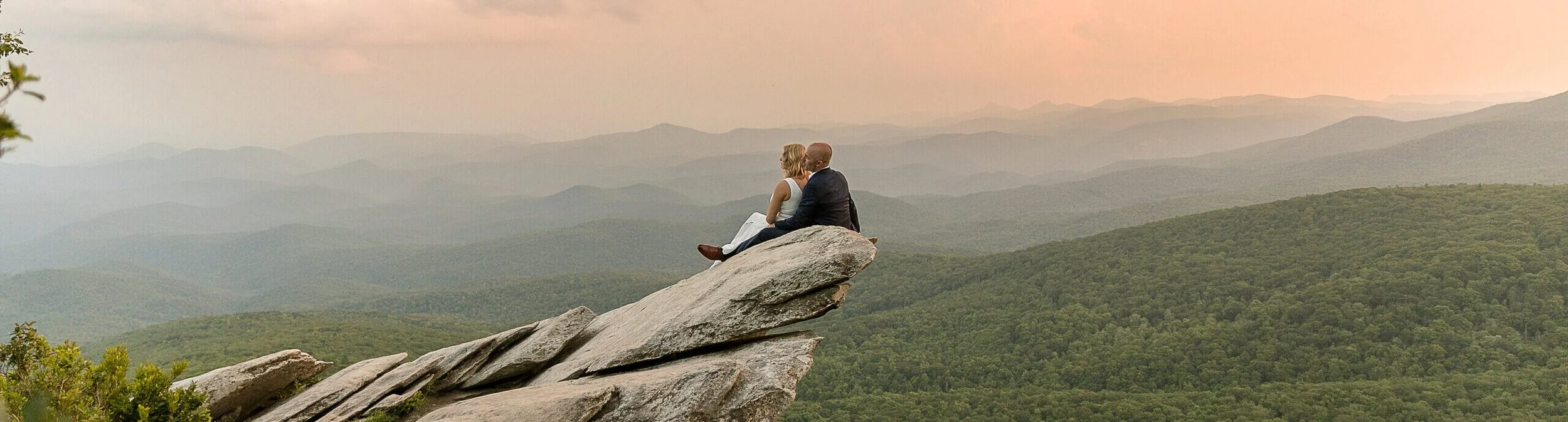 A couple sitting on a large rock formation at the edge of a cliff overlooking a mountain landscape at sunset.