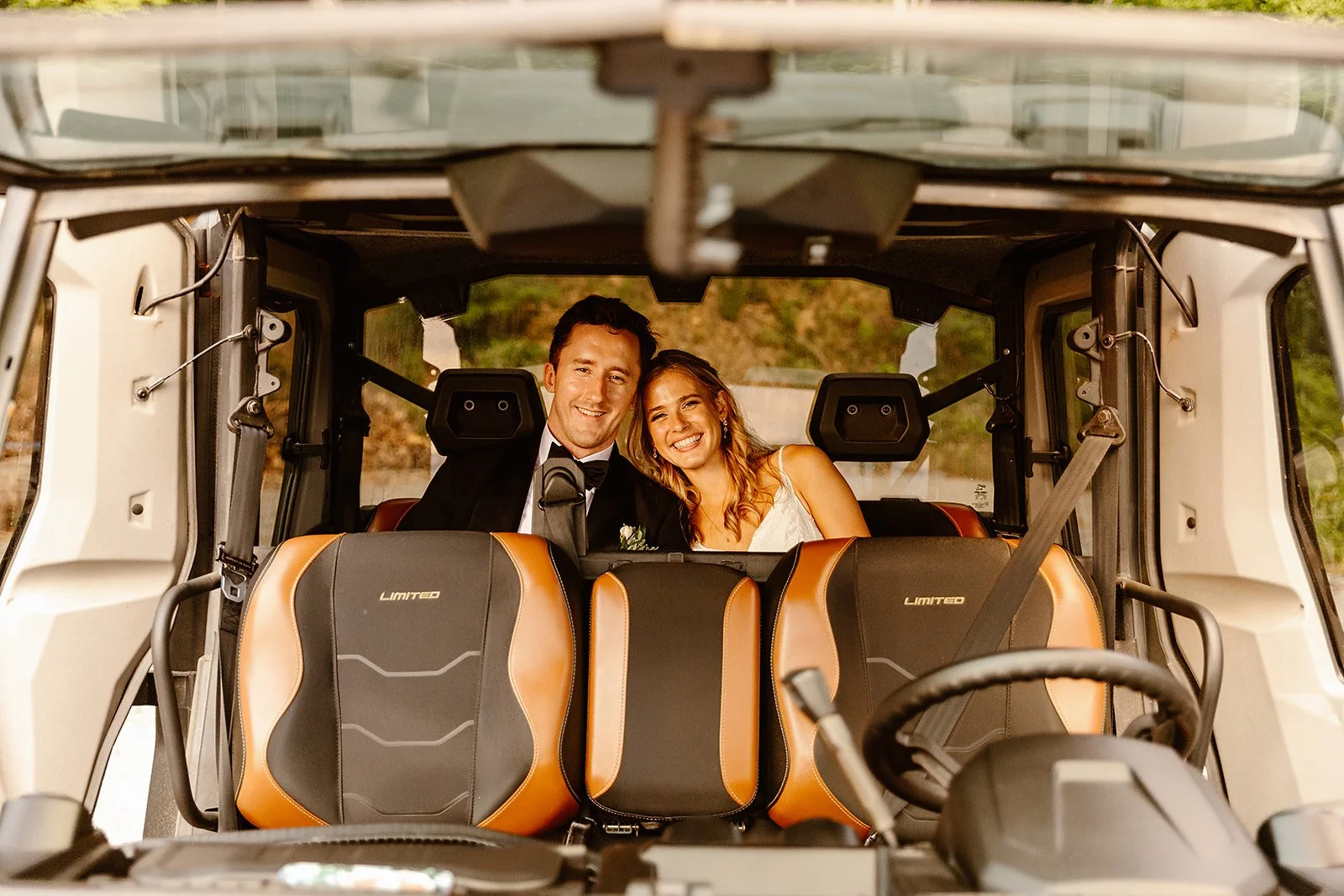 A man and woman sitting inside a golf cart, dressed in formal wedding attire, smiling at the camera.