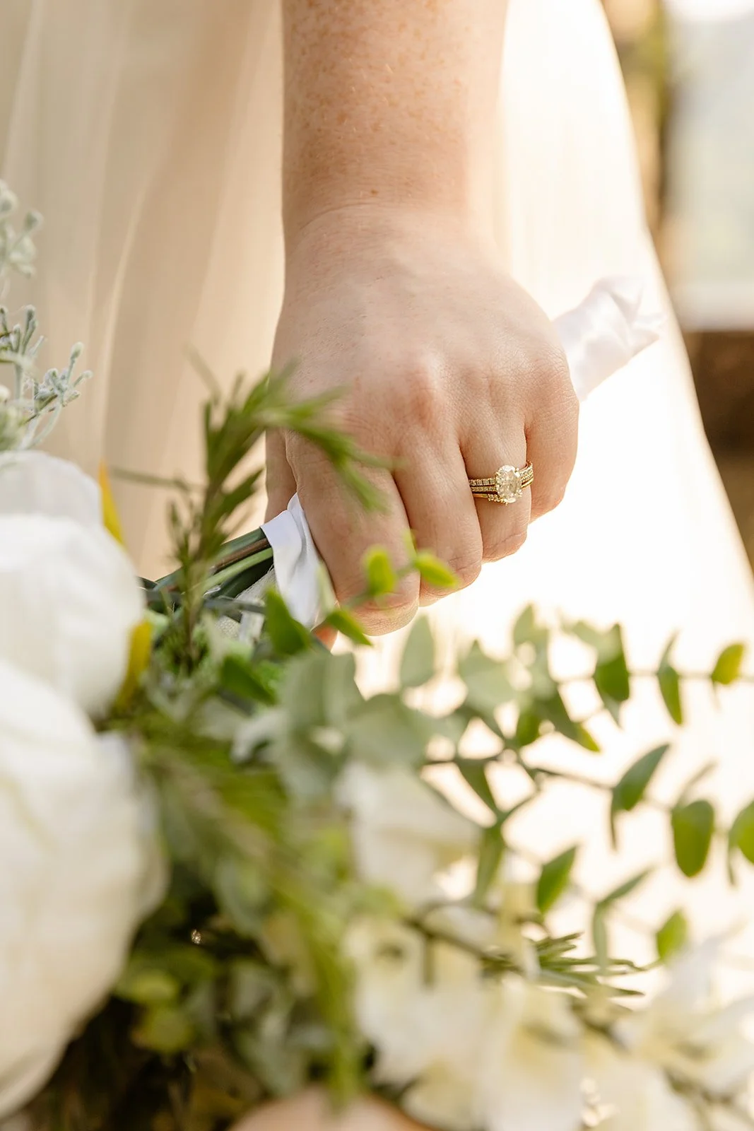 Close-up of a woman's hand with a gold wedding ring, holding a bouquet of white flowers and green foliage.