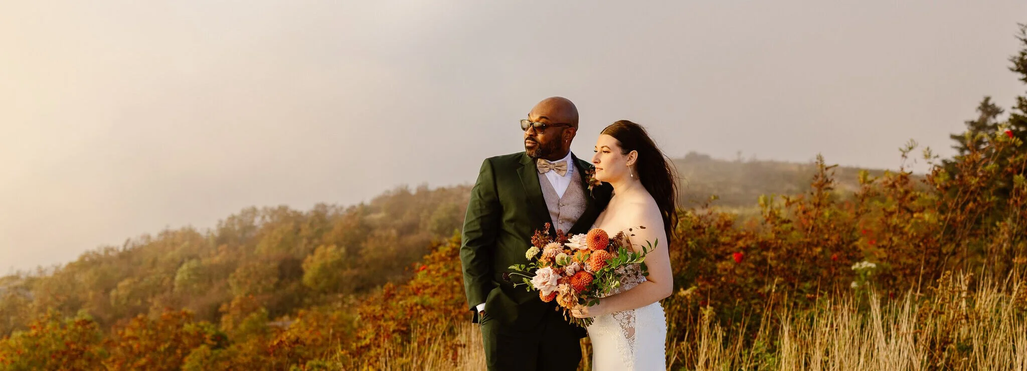 A bride and groom standing together outdoors during sunset on a grassy hill, with trees in the background. The bride is holding a large bouquet of colorful flowers, and the groom is dressed in a dark suit with glasses and a bow tie. Both are looking pensively to the side.