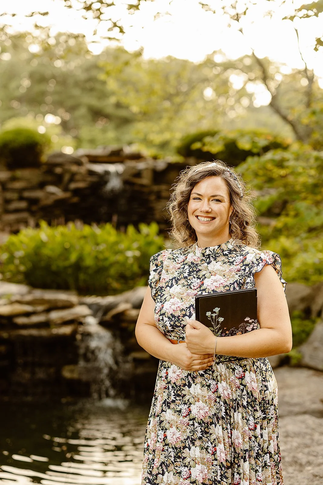 A woman with curly hair smiling outdoors, holding a black notebook with floral design, standing near a small waterfall in a garden.