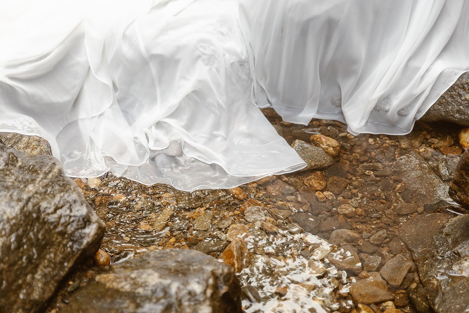 A white wedding dress submerged in a shallow rocky stream of water.