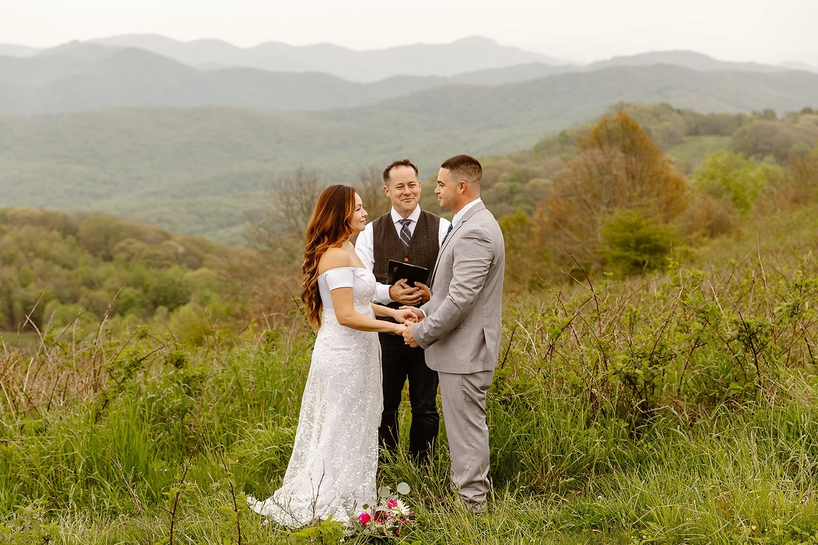A couple getting married outdoors during daytime, holding hands and looking at each other, with an officiant standing behind holding a book, in a grassy field with mountains and trees in the background.
