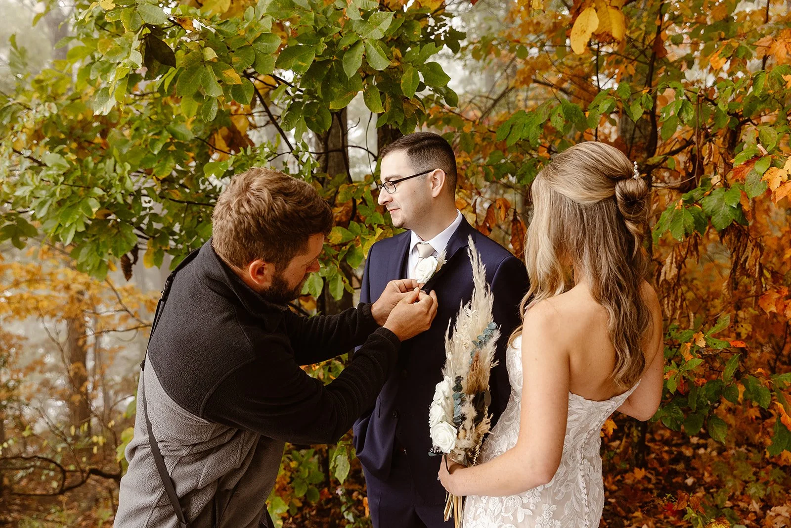A wedding ceremony outdoors in autumn, with a bride and groom standing among colorful fall foliage, as a man is pinning a boutonniere onto the groom's suit.