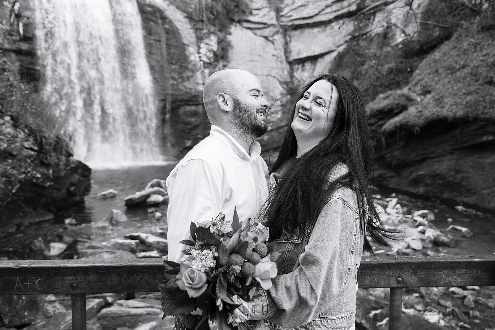 A happy couple in love, standing in front of a waterfall, with the woman holding a bouquet of flowers, both smiling and laughing.