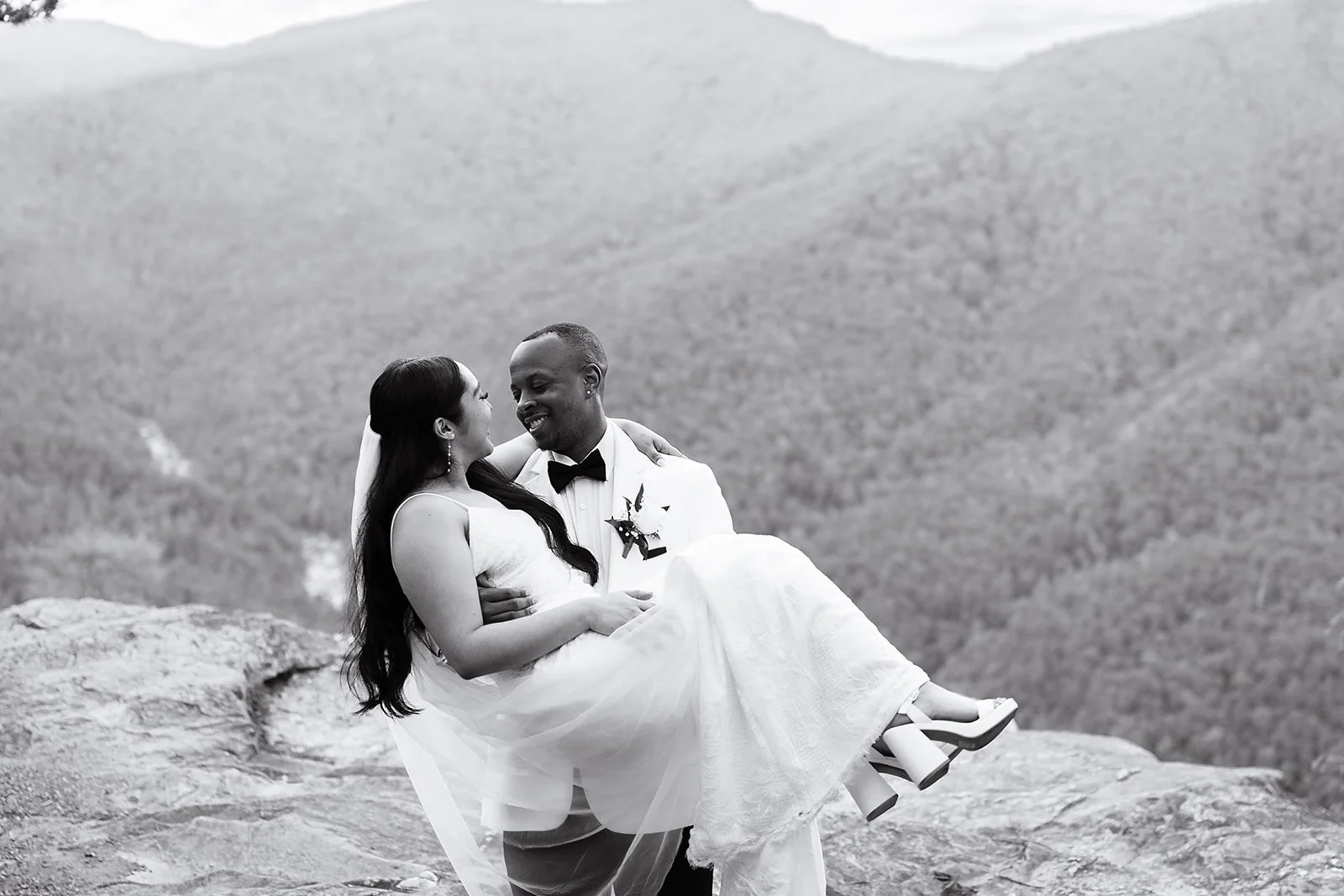 A black-and-white photo of a bride and groom, with the groom holding the bride in his arms, sitting on a rocky ledge with mountain terrain in the background, smiling at each other.