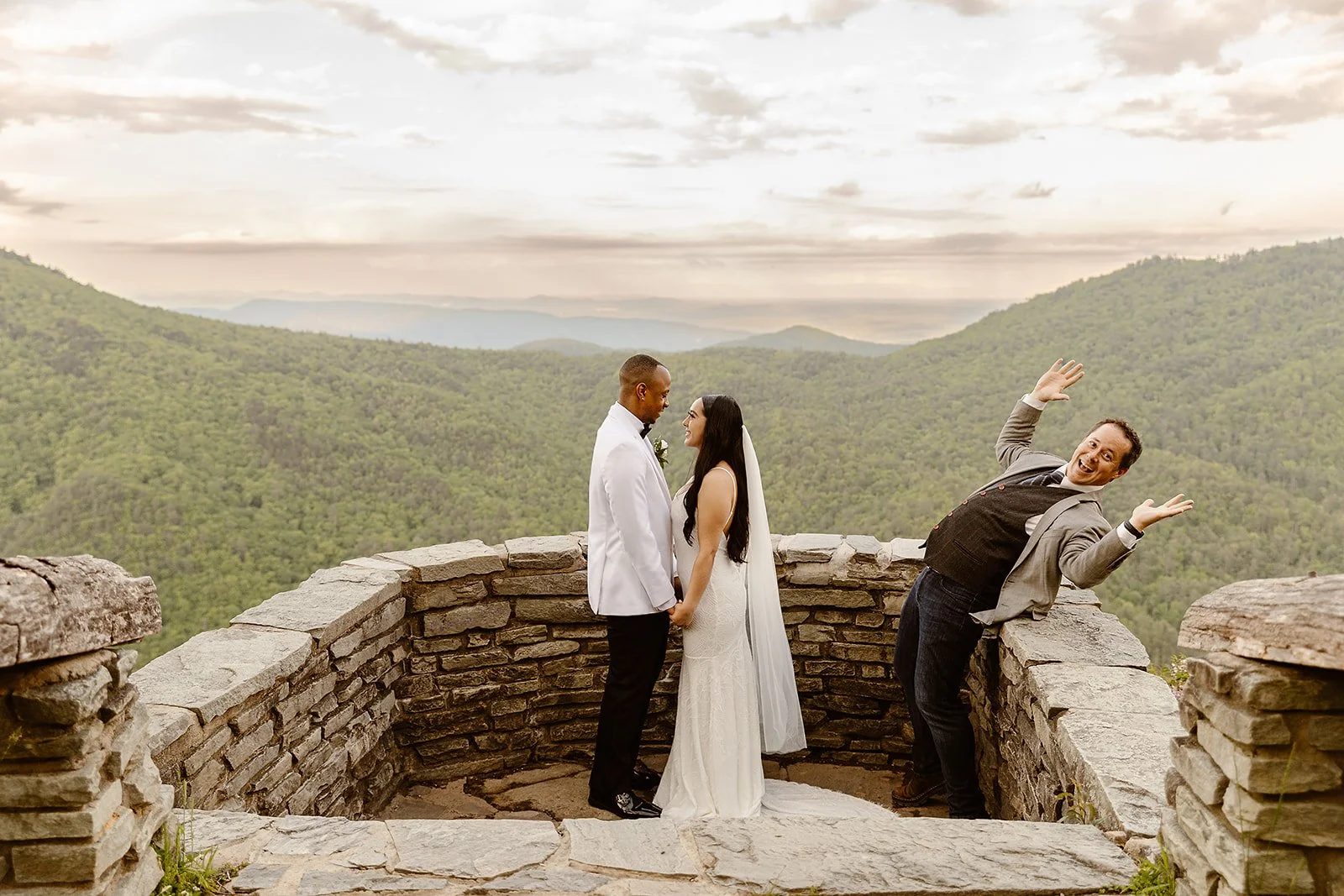 A newlywed couple holding hands and smiling at each other on a stone lookout with mountains in the background, while another man leans back and gestures playfully.
