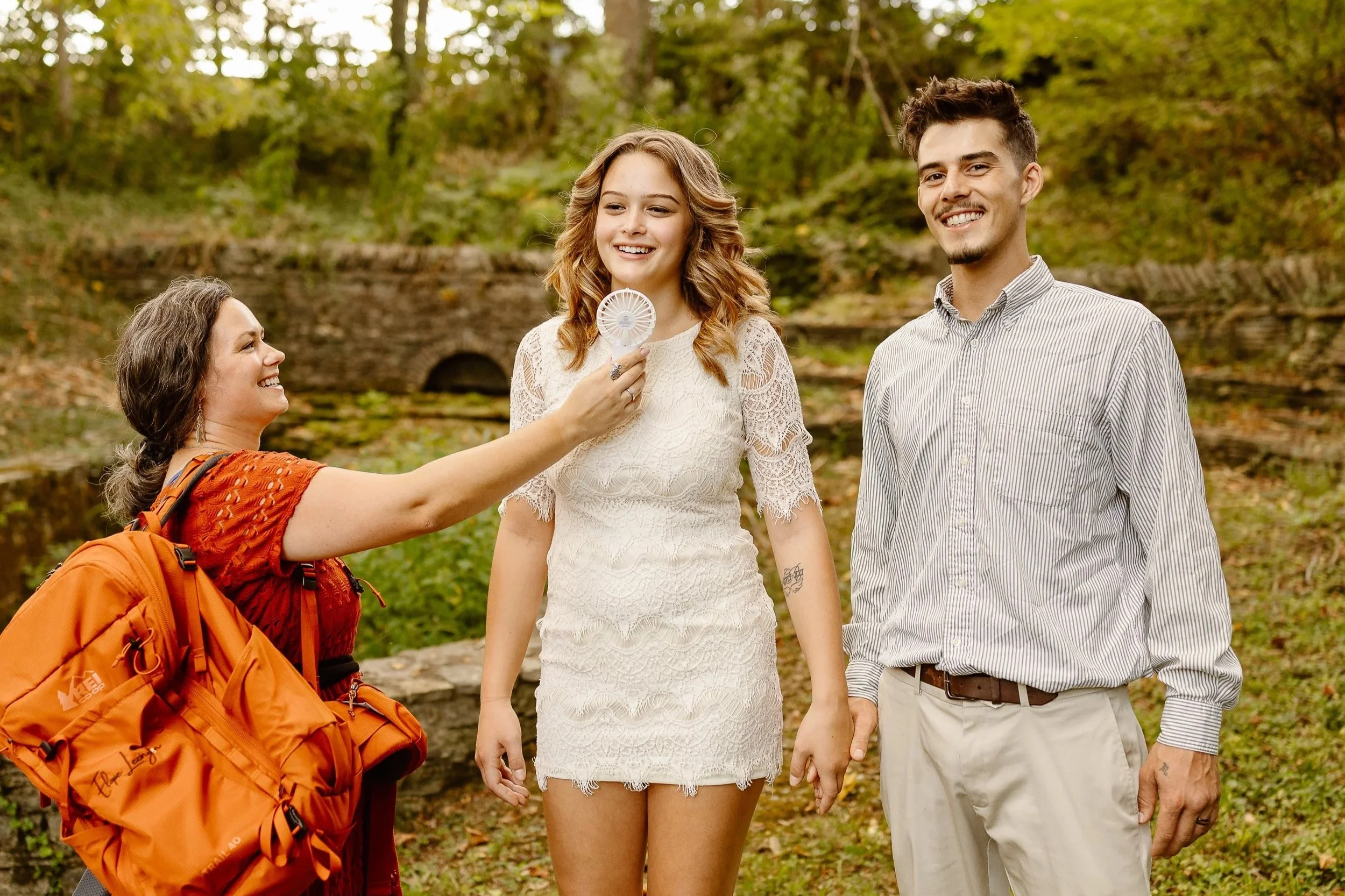 Five people outdoors in autumn, a woman with a backpack is holding a small fan near a young woman's face, who is smiling and wearing a white lace dress, while a young man in a striped shirt smiles nearby. There is a stone bridge and fall foliage in t
