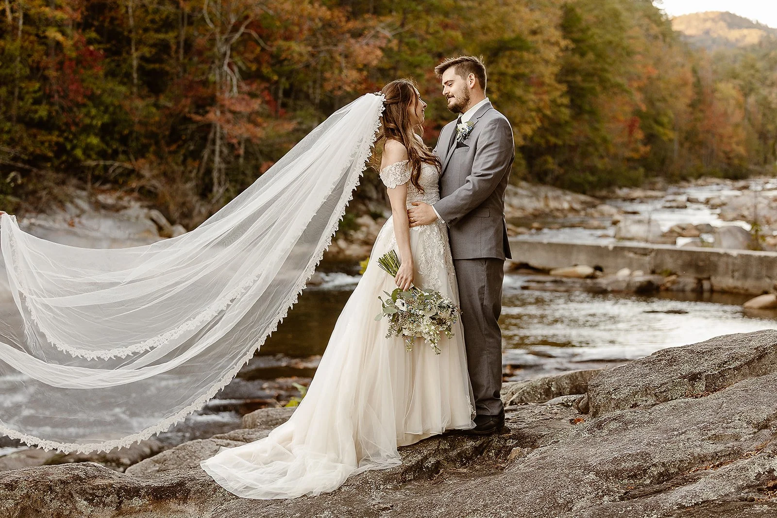 A bride and groom standing on rocks by a river, facing each other in wedding attire, with a background of autumn trees and a flowing river.