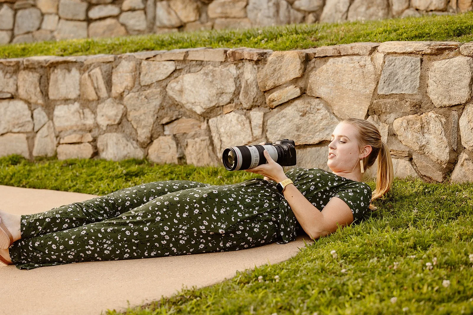 A young woman lying on the grass next to a stone wall, holding a professional camera with a large telephoto lens, appearing to be taking photos outdoors.