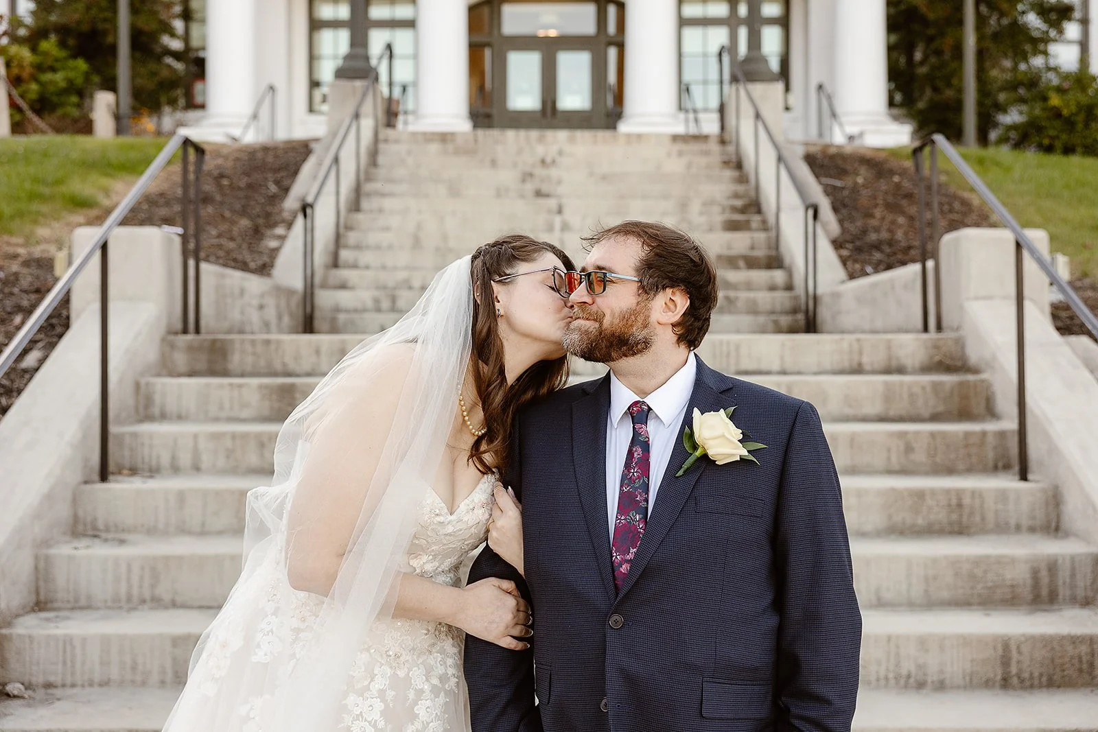 A bride and groom kissing on the steps of a building, with the bride wearing a wedding dress and veil, and the groom wearing a suit with a boutonnière.