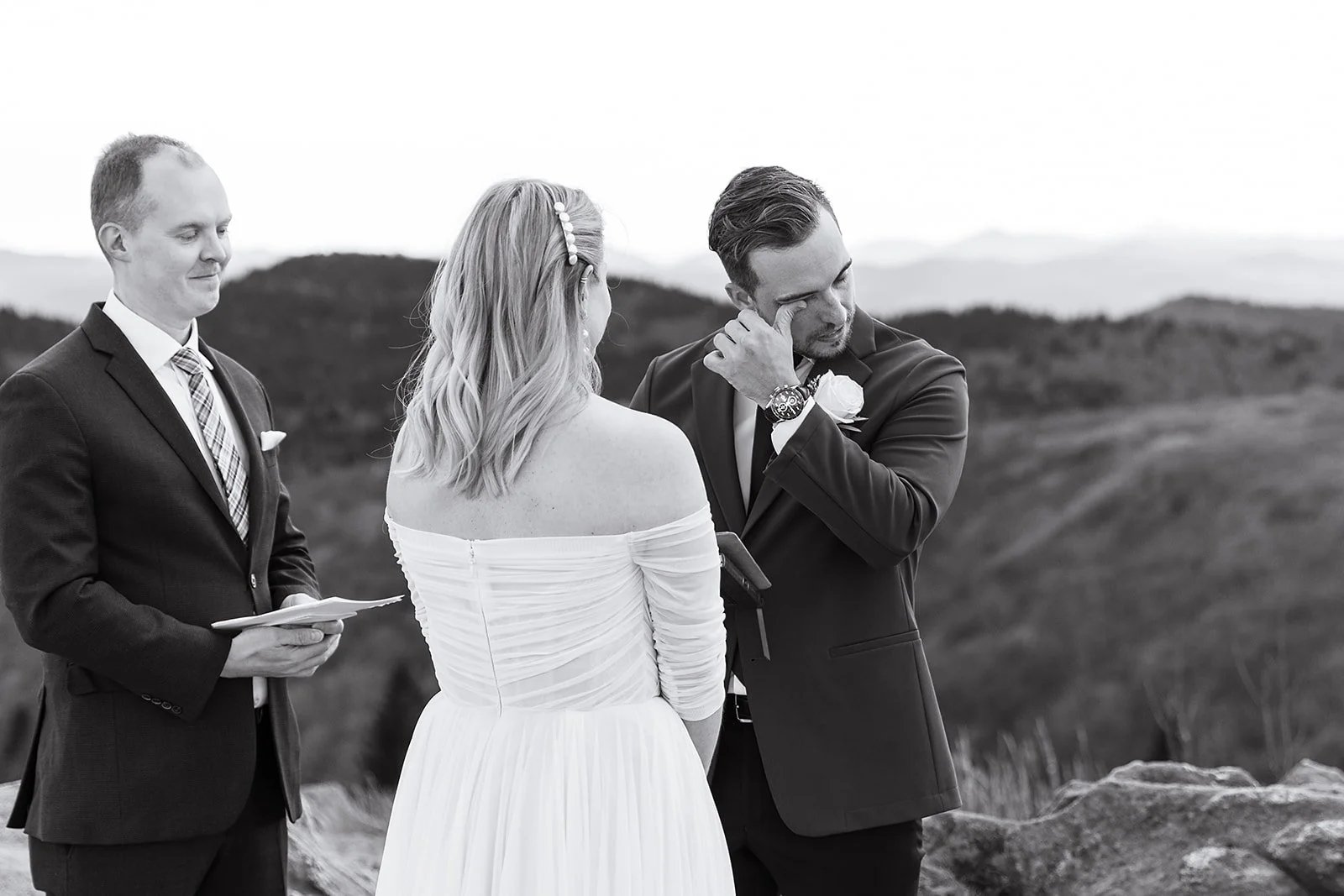 A black-and-white photo of a wedding ceremony outdoors with a bride, groom, and officiant, with mountains in the background.