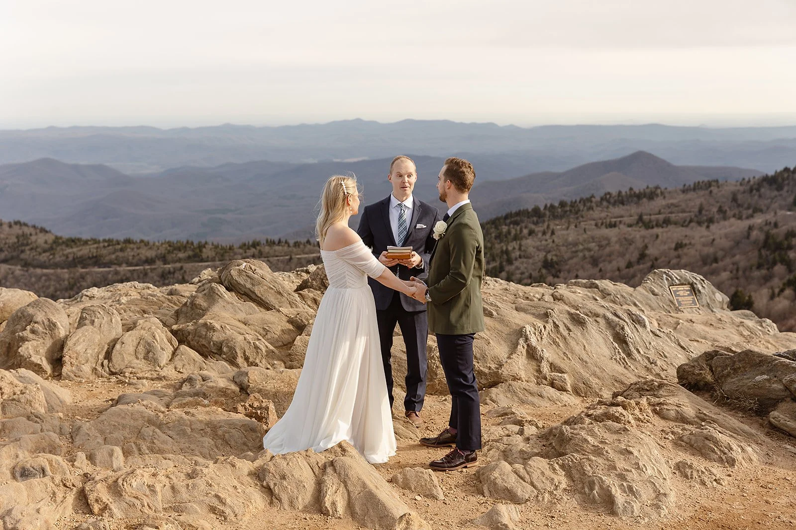 A couple getting married outdoors on rocky terrain with mountains in the background, holding hands and facing each other, with a officiant present.