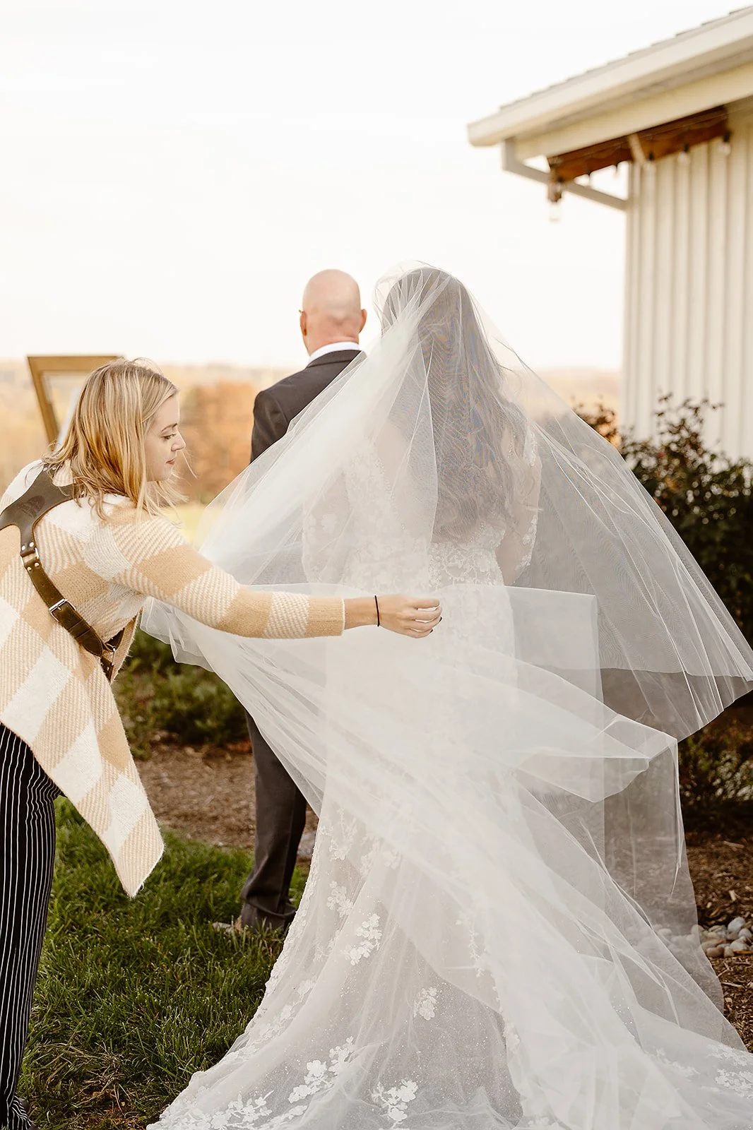 A woman helping a bride set up her veil outdoors during a wedding ceremony, with a man standing nearby and a white building in the background.