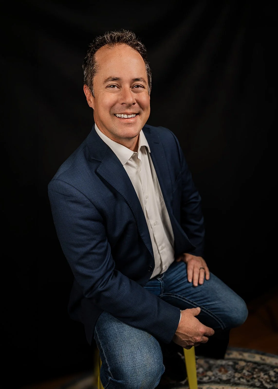 A smiling man with short brown hair, wearing a navy blazer, white dress shirt, and blue jeans, sitting on a yellow stool against a black background.