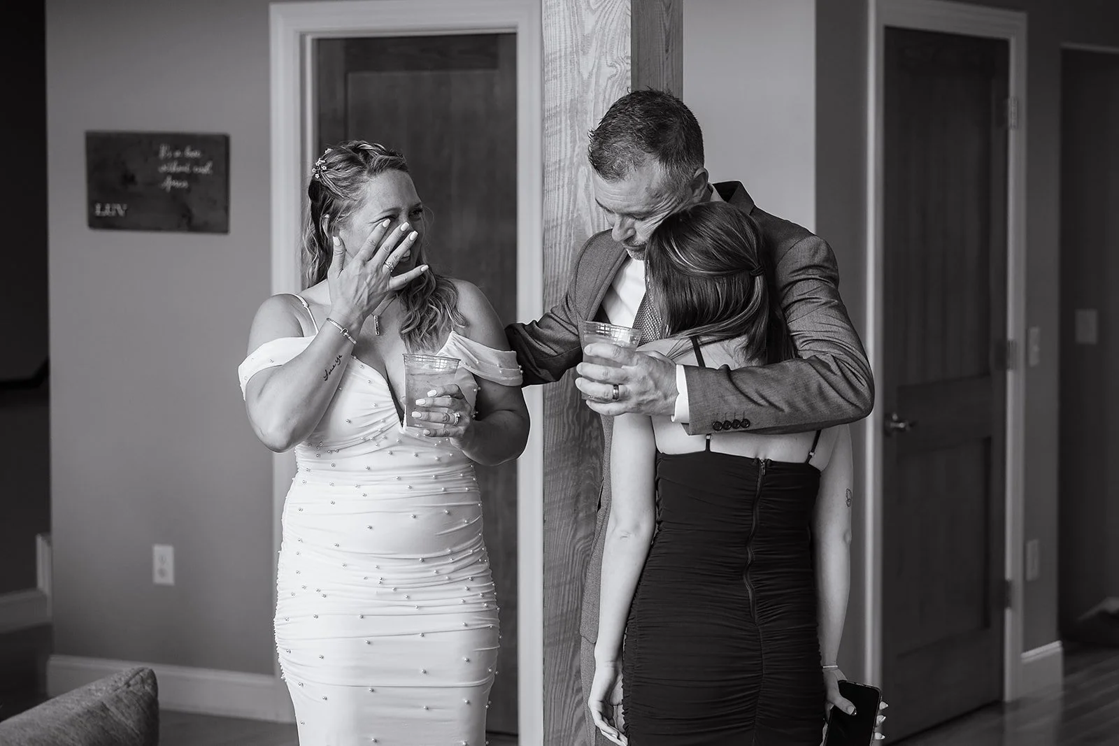 Three people share an emotional moment indoors, two women and one man, with the woman on the left wearing an elegant dress, the man in a suit, and the woman on the right in a black dress, holding drinks.