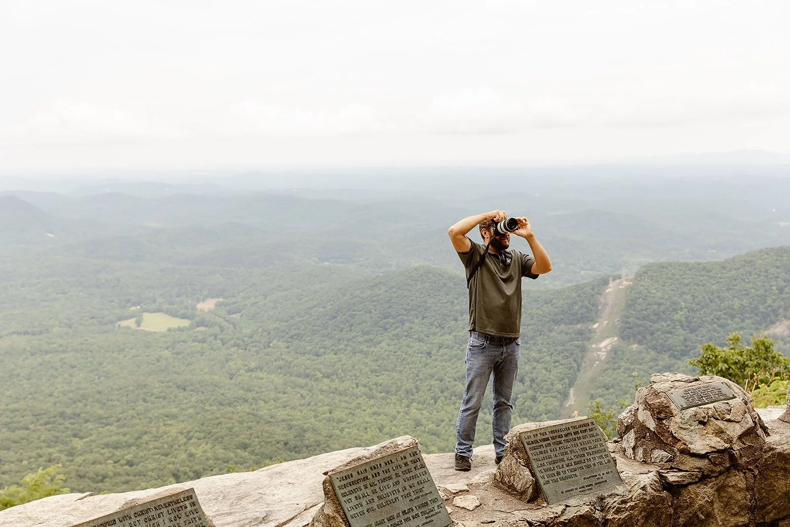 A man standing on a rocky ledge, taking a photo with a camera, overlooking a vast green landscape with hills and forests under a cloudy sky.