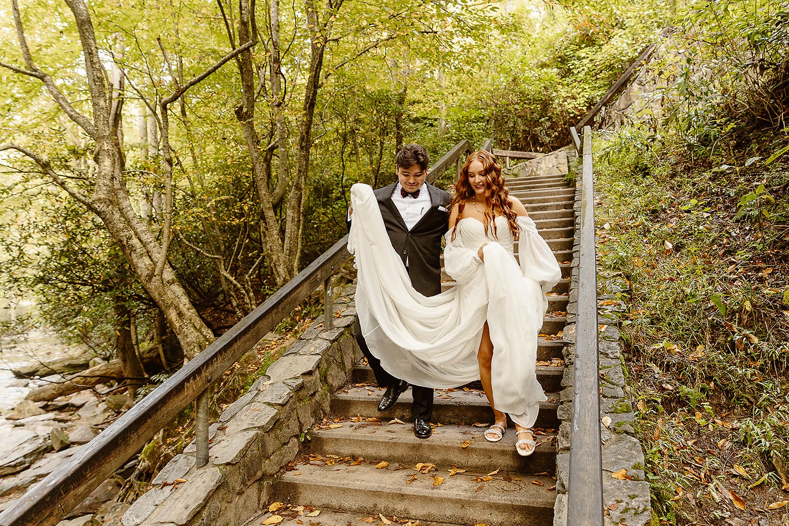 A bride and groom walking down outdoor stone stairs surrounded by trees and greenery.