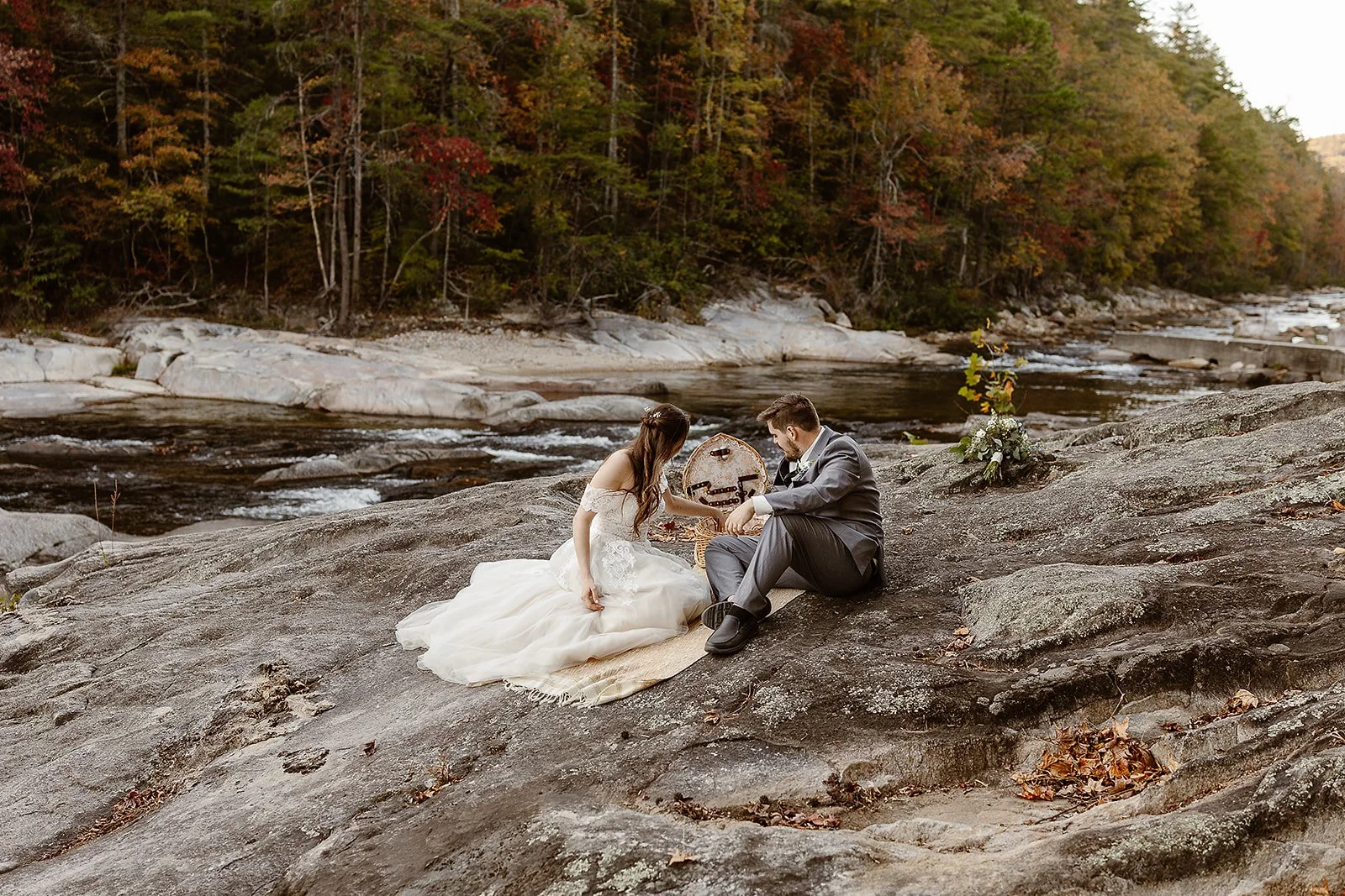 A bride and groom sitting on a large rock by a river, holding hands during their wedding ceremony, with trees displaying fall colors in the background.