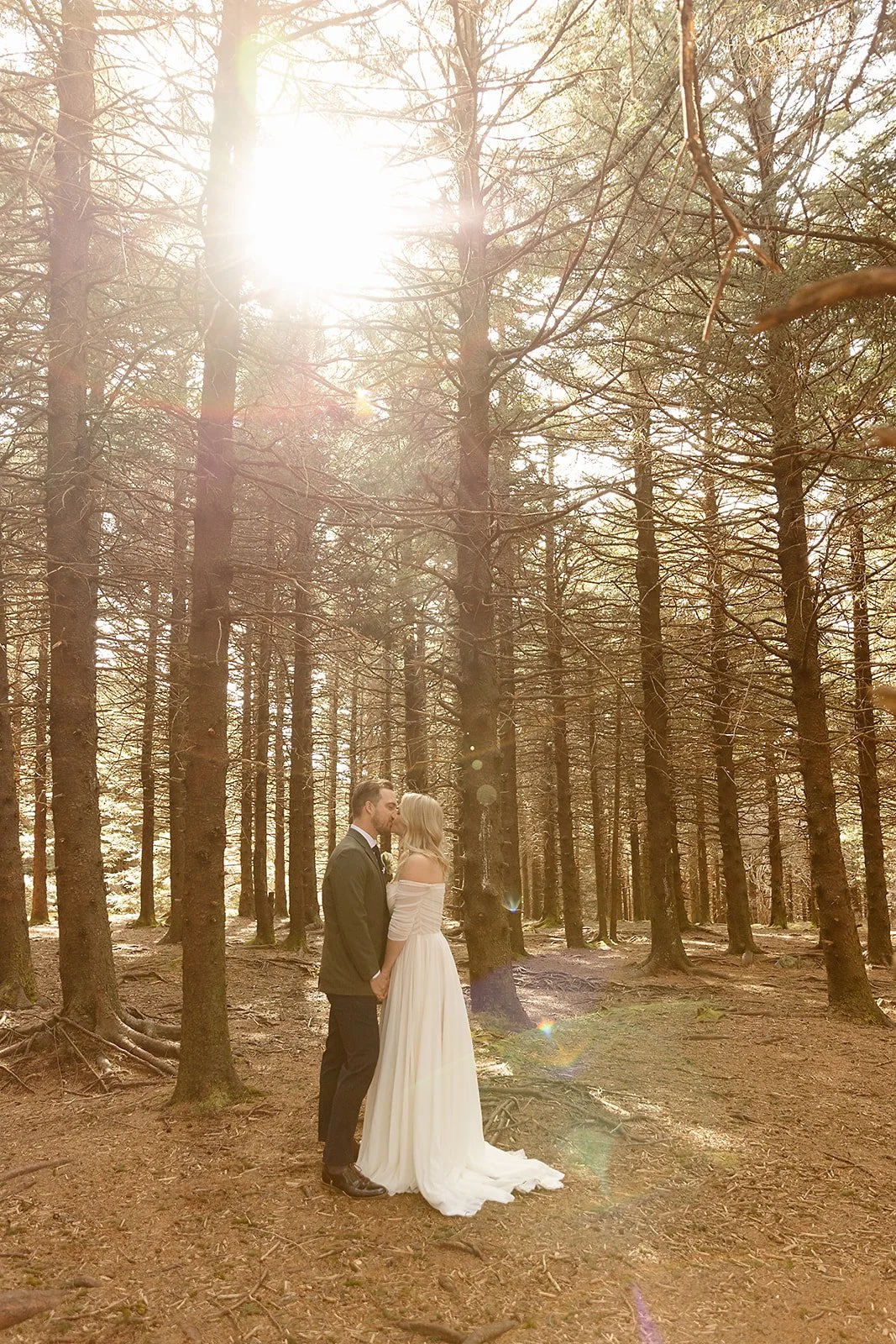 A couple dressed in wedding attire sharing a kiss in a sunlit forest with tall trees and ground covered in leaves and roots.