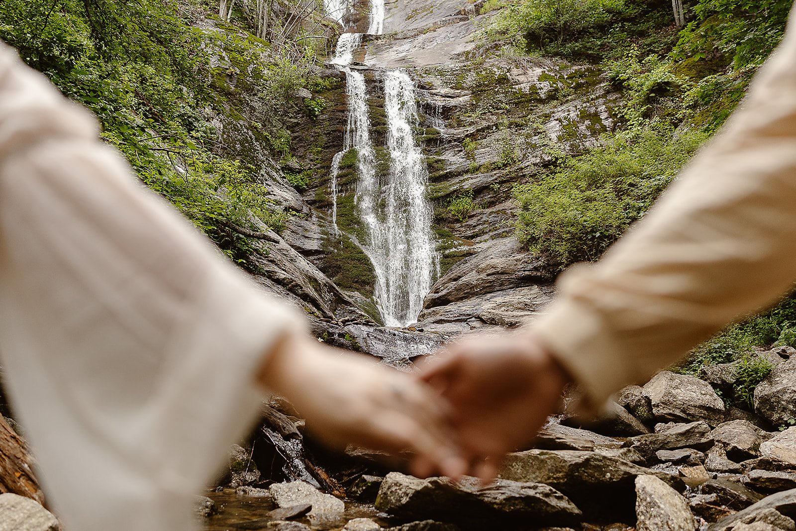 A couple holding hands in front of a waterfall in a forest.