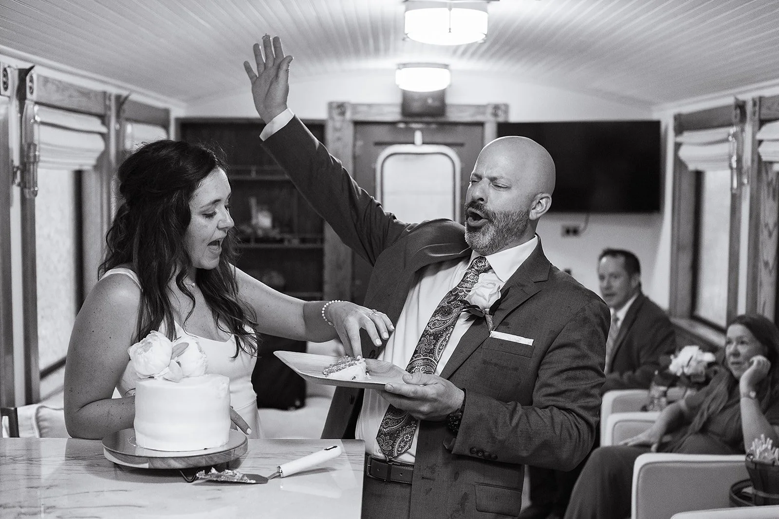 A black and white photo of a wedding reception with a bride, groom, and guests. The groom is singing or speaking passionately, holding a serving tray, with his right arm raised. The bride has her eyes closed and is touching the tray. Guests are seated in the background, watching the scene.