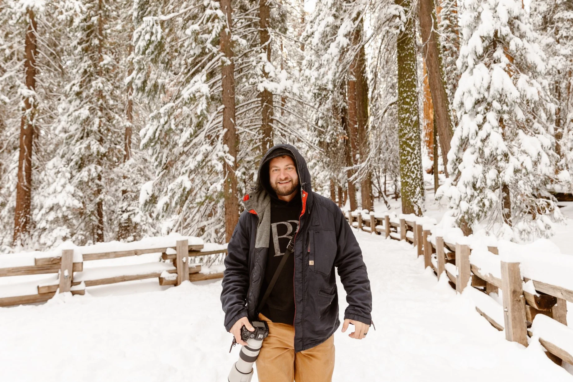 A man in a black jacket and tan pants standing in a snowy forest with evergreen trees, holding a camera with a large lens.