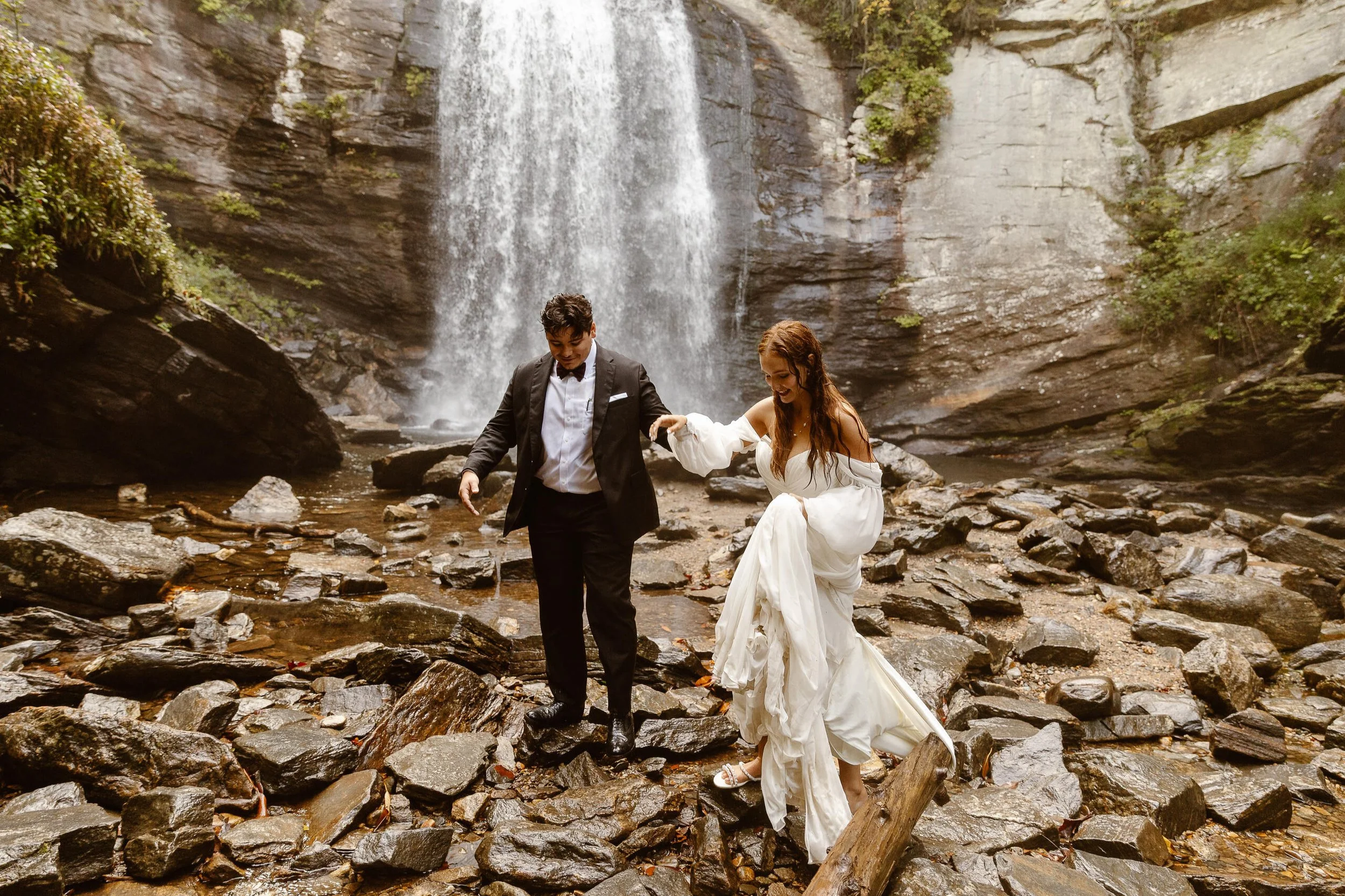 A bride and groom in wedding attire walking on rocky terrain near a waterfall in a forested area.