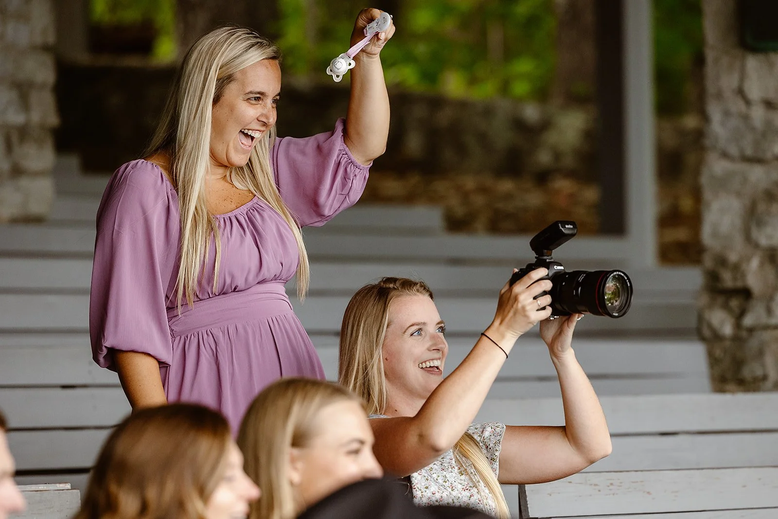 A woman in a lavender dress is standing and smiling while holding a pair of 3D glasses. Another woman sitting in front is smiling as she takes a photo with a camera. Other people are partially visible in the foreground, sitting in an outdoor setting 