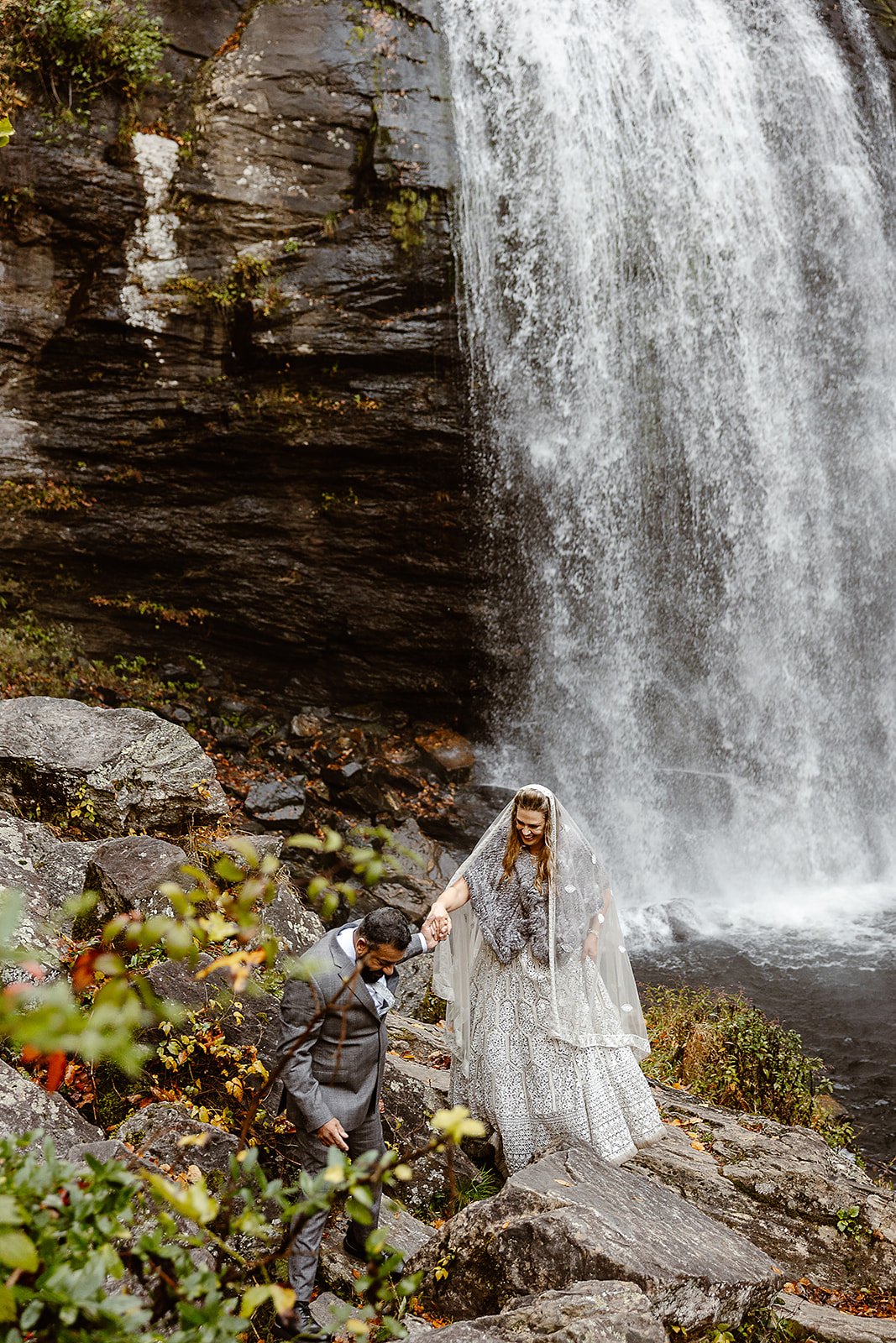 Bride and groom in wedding attire standing near a waterfall, holding hands on rocky terrain.