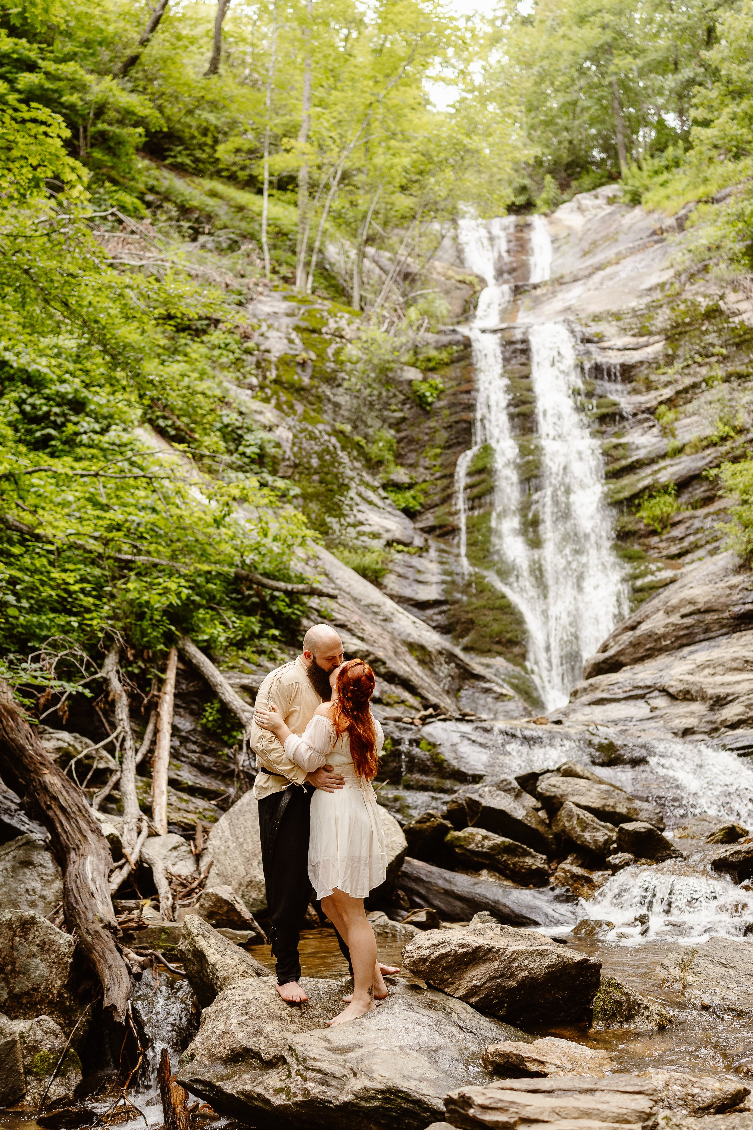 A couple embracing on rocks in front of a waterfall in a lush green forest.