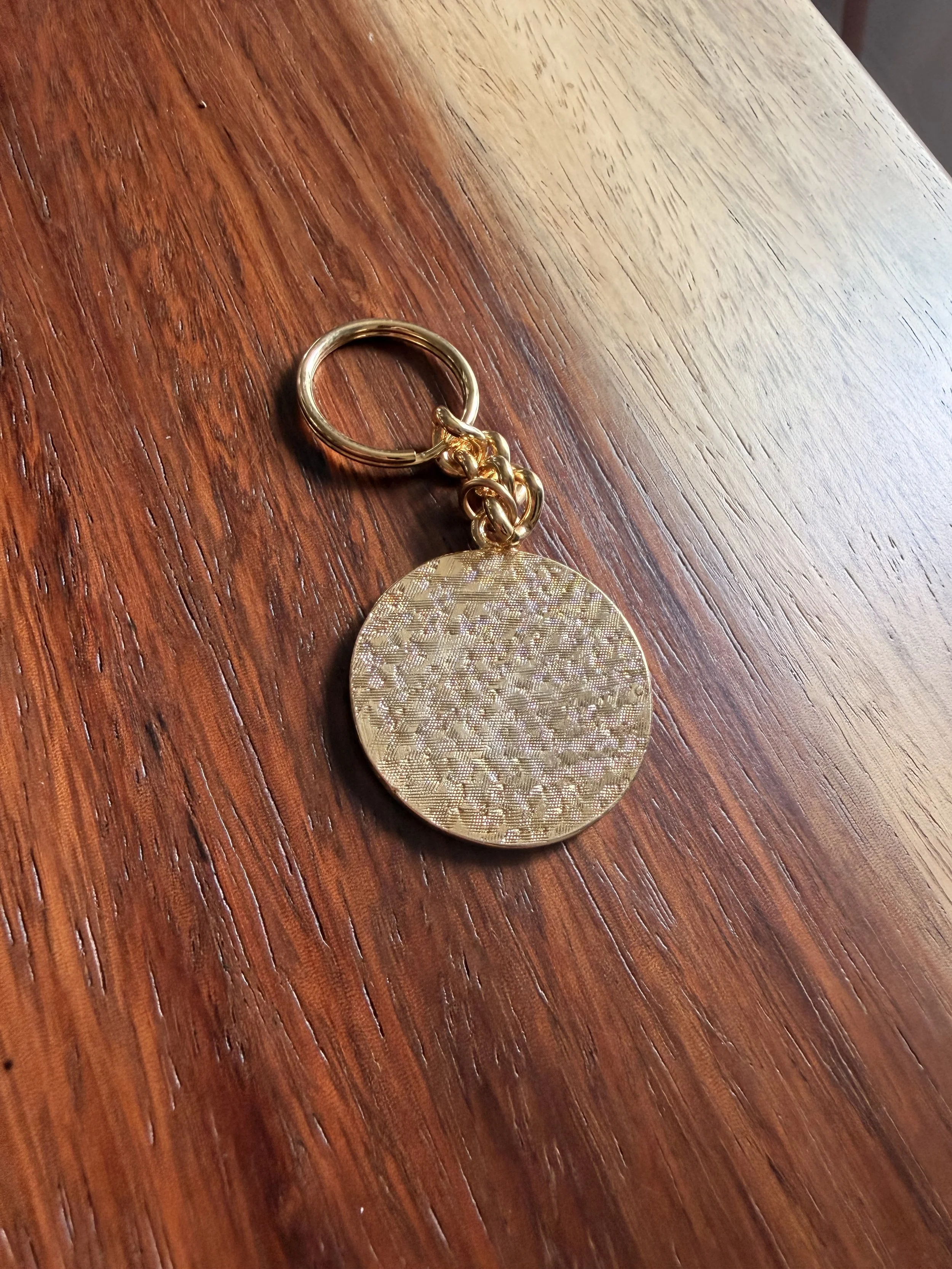 A gold-colored keychain with a large, round, textured pendant lying on a wooden surface.