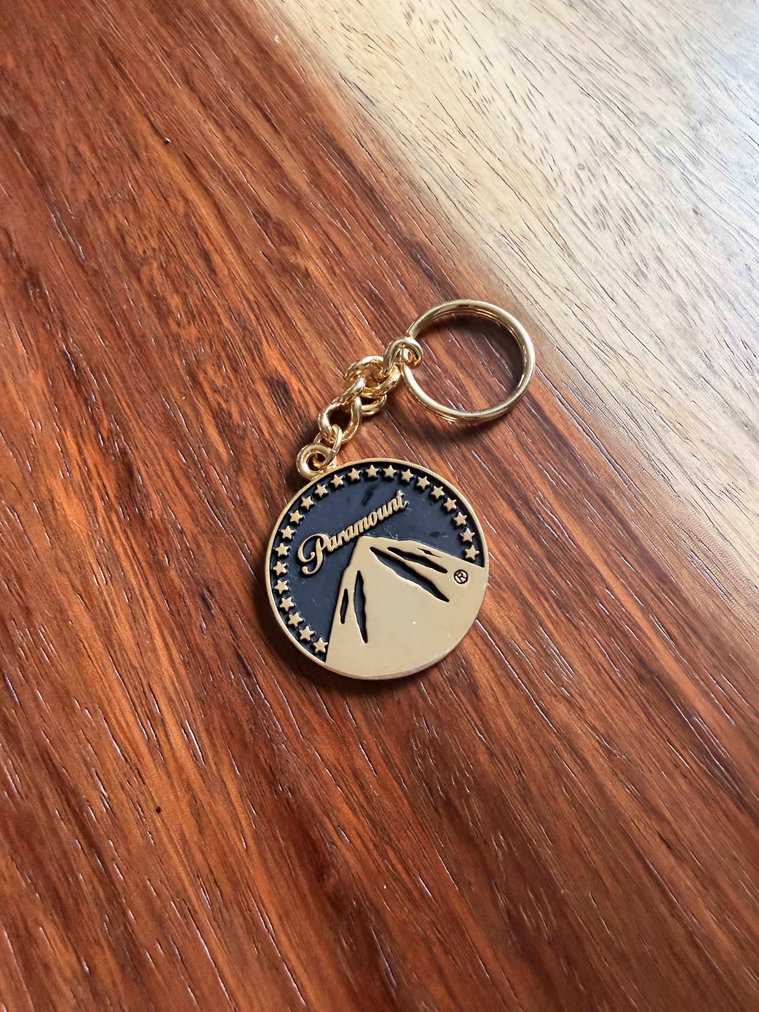 A gold-colored keychain with the Paramount Pictures logo, featuring a stylized mountain and stars, resting on a wooden surface.