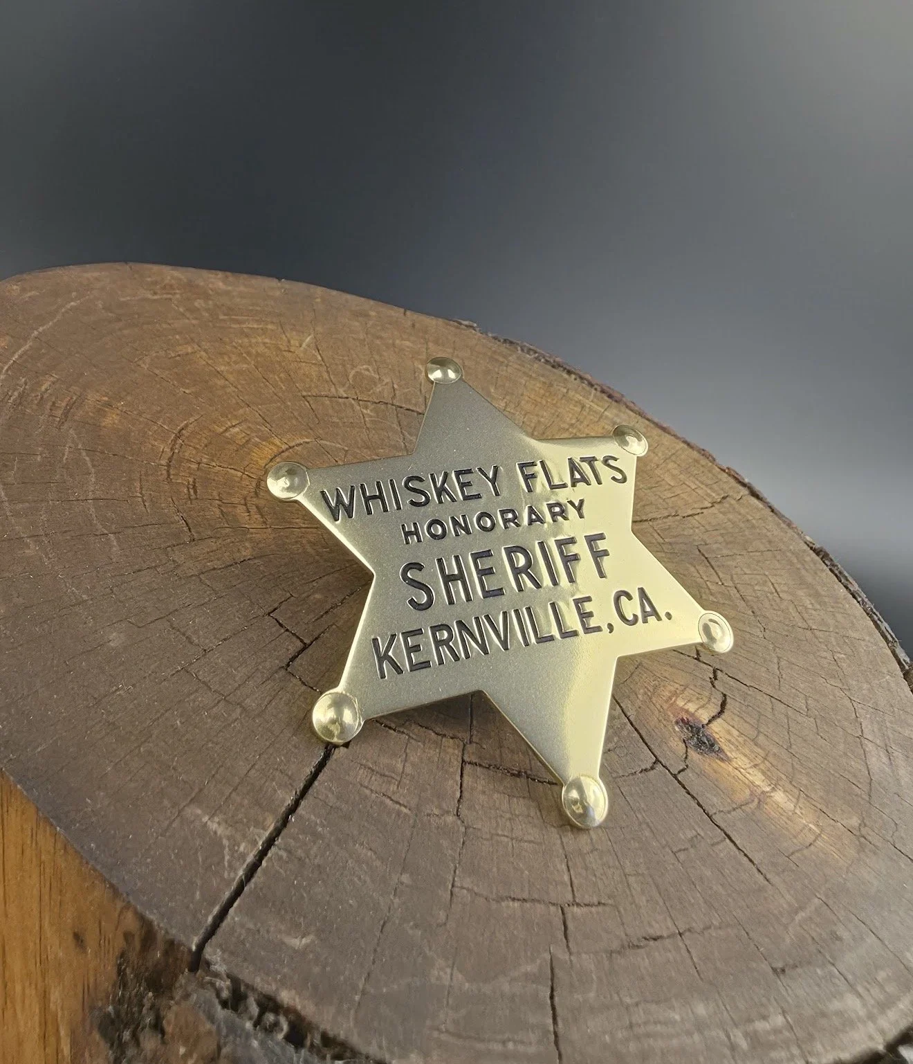 A sheriff's badge mounted on a wooden log with visible cracks and rings, reading 'Whiskey Flats Honorary Sheriff Kernville, CA'.