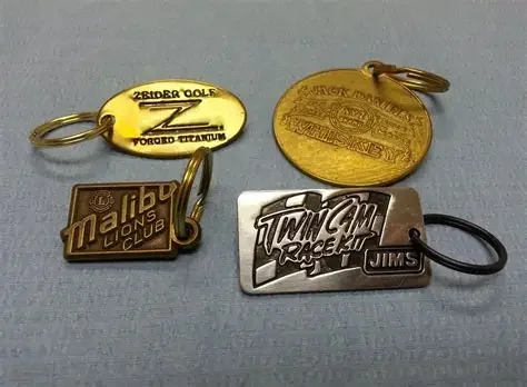 Four keychains, including one yellow oval with 'Zeider Golf' and 'Torded Titanium,' one gold oval with engraved text, a brown rectangular 'Mild Two Coins Club' keychain, and a silver 'Twin Cam Race Kit Jims' keychain, all on a gray surface.