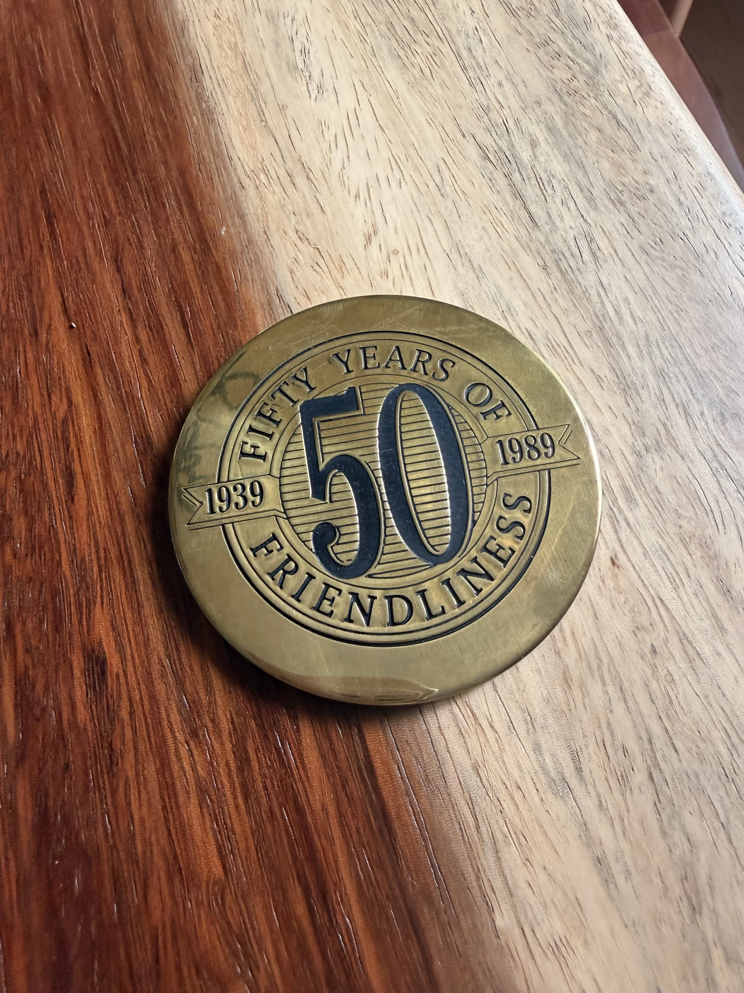 A gold-colored round medal with the number 50 in the center, surrounded by the inscription 'FIFTY YEARS OF FRIENDLINESS' and the dates '1939' and '1989', placed on a wooden surface.