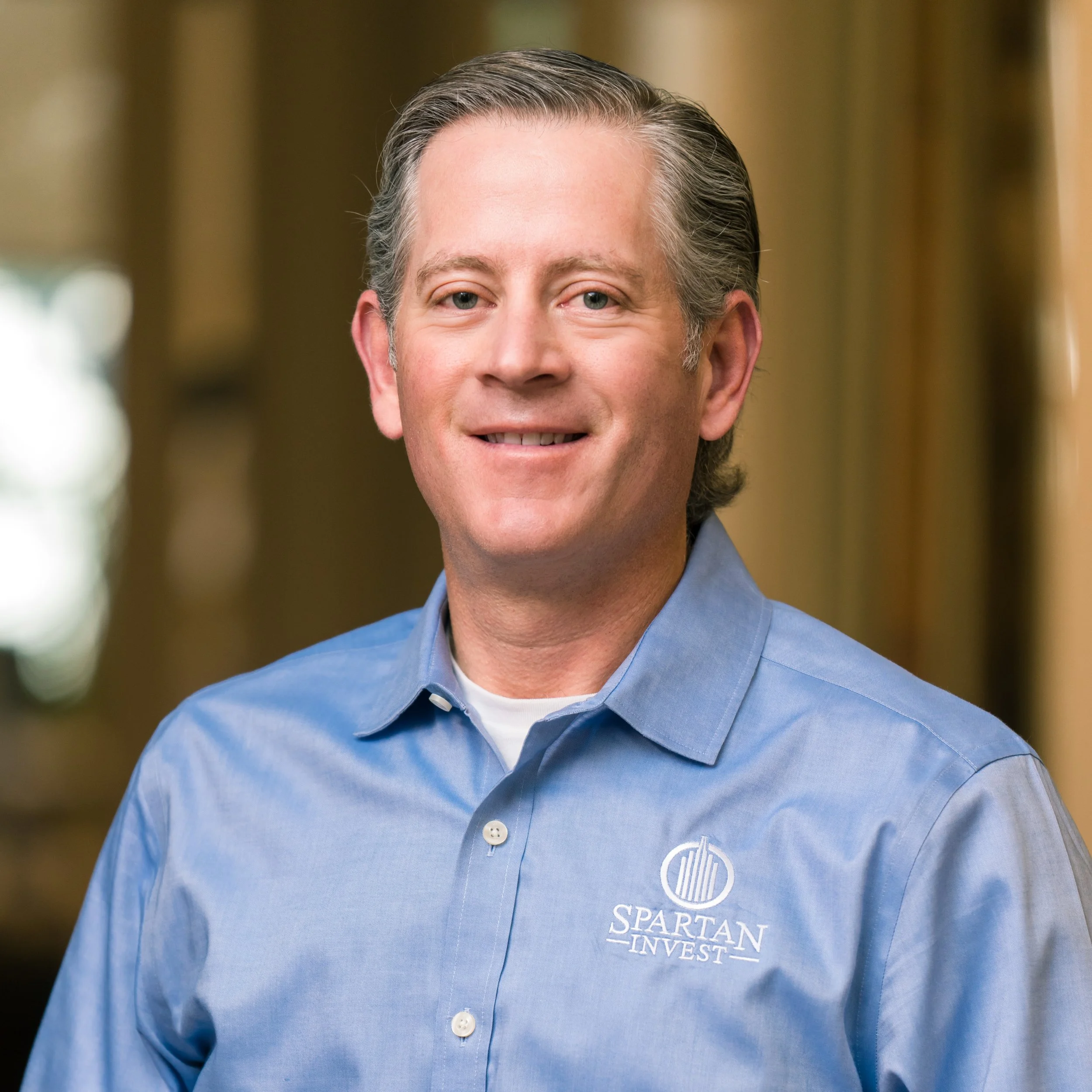 Close-up of a man wearing a blue collared shirt with the logo 'Spartan Invest' visible on the pocket area.