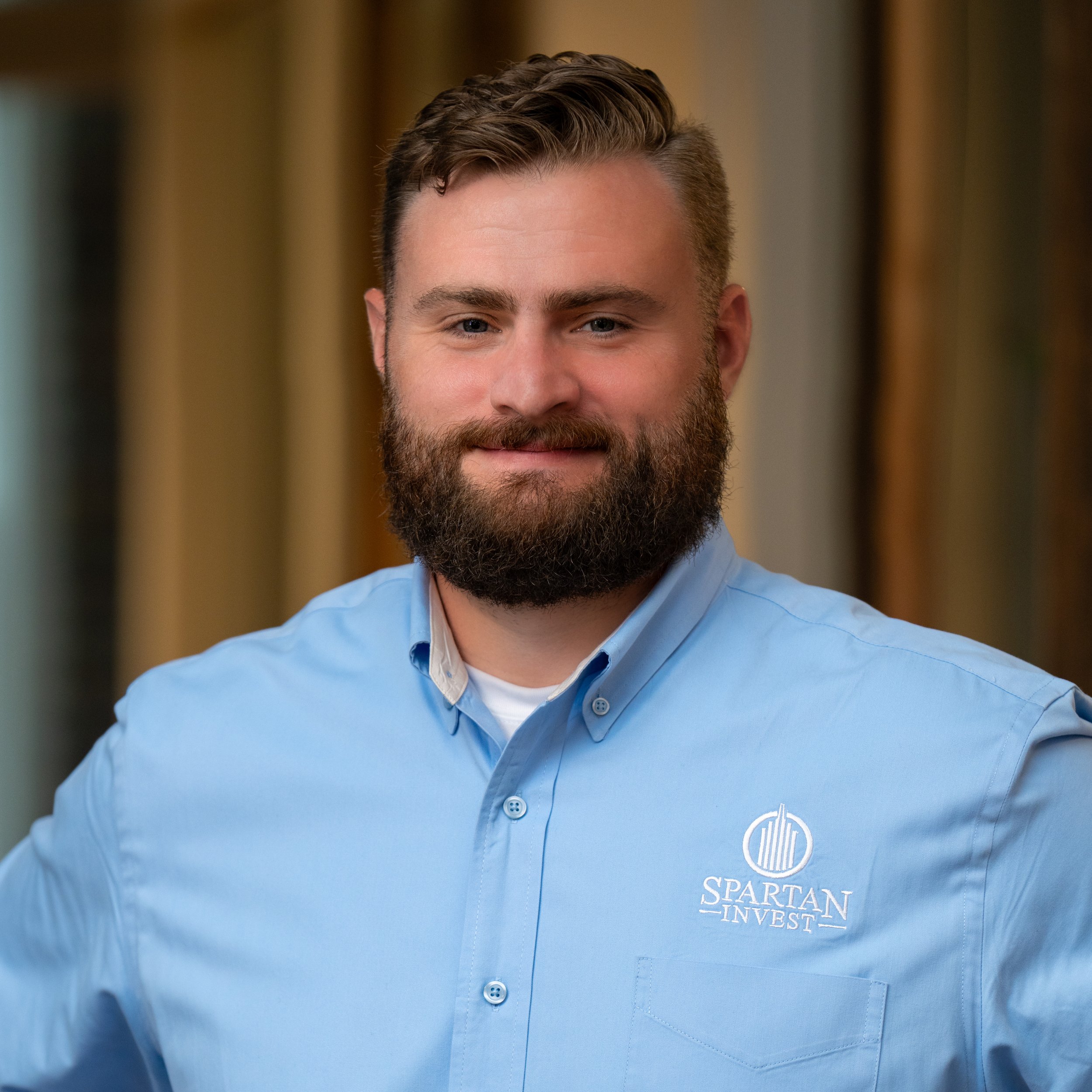Close-up of a man's face with a beard, wearing a light blue shirt with a 'Spartan Invest' logo on the pocket, indoors with blurred background.
