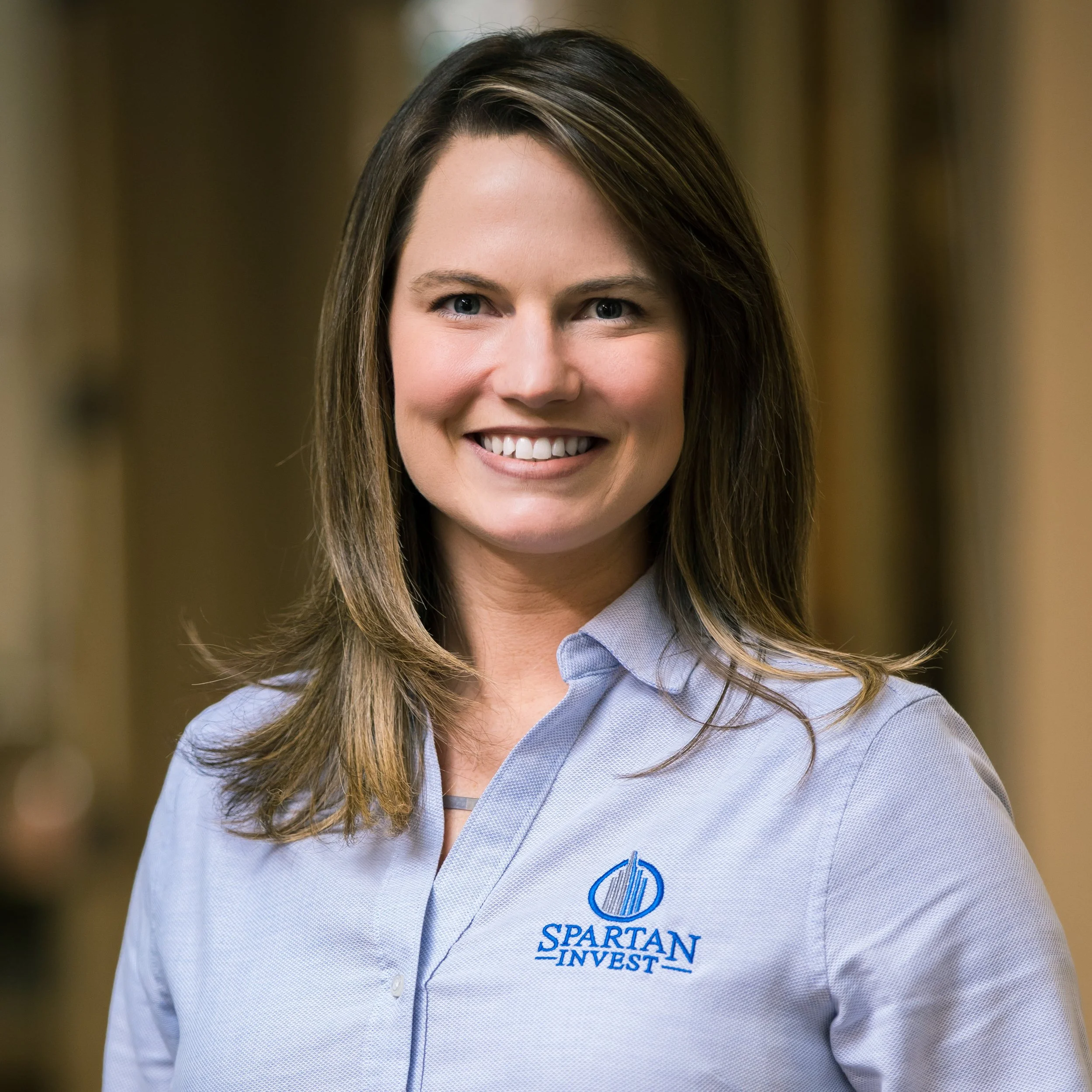Close-up of a woman with shoulder-length brown hair, blue eyes, smiling, wearing a light blue collared shirt with the logo and text 'Spartan Invest' on it.