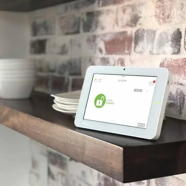 Smart home control panel on a dark wooden shelf with plates stacked nearby against a brick wall.