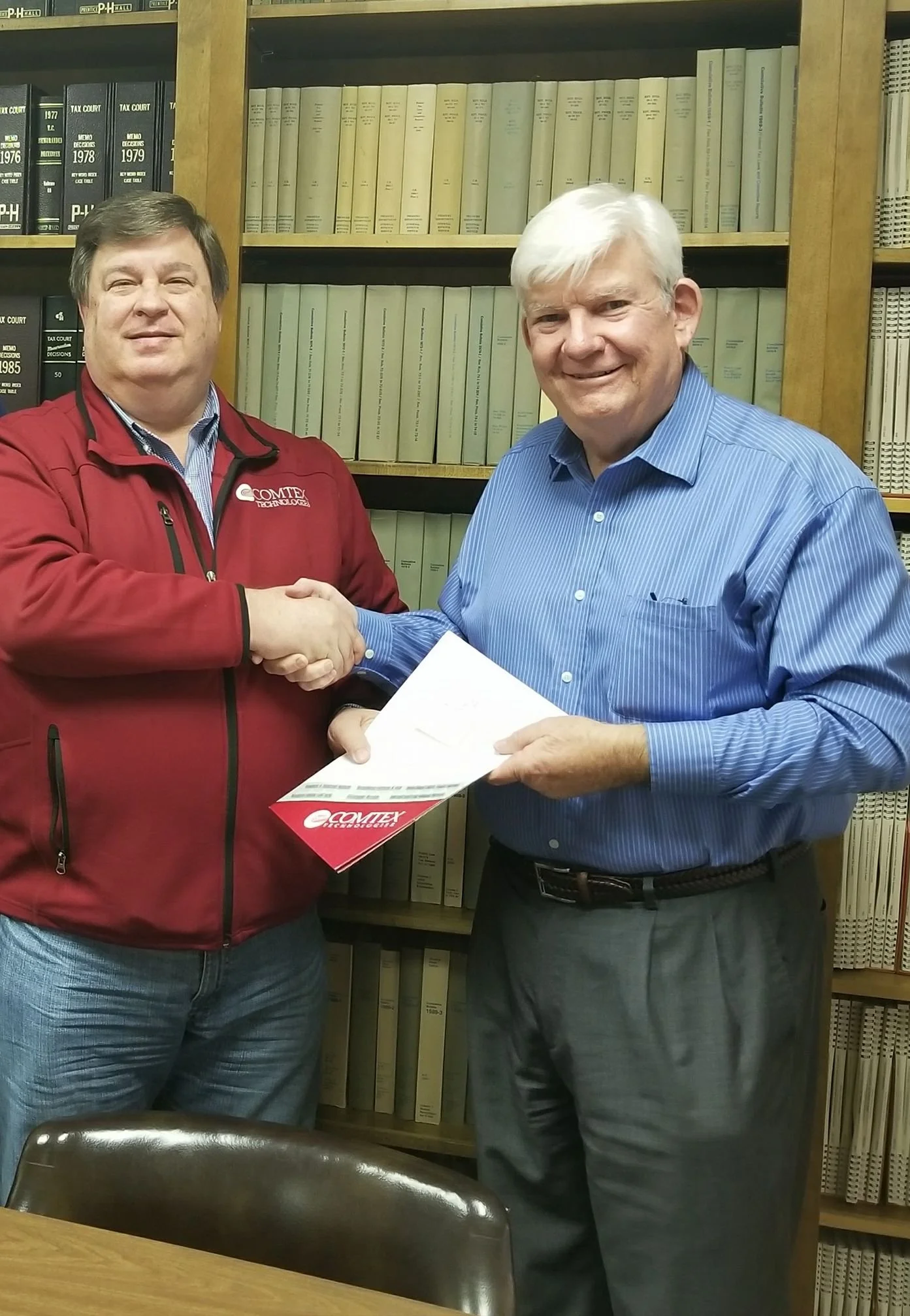 Two men shaking hands in a library or office, one wearing a red jacket and the other wearing a blue striped shirt, both smiling.