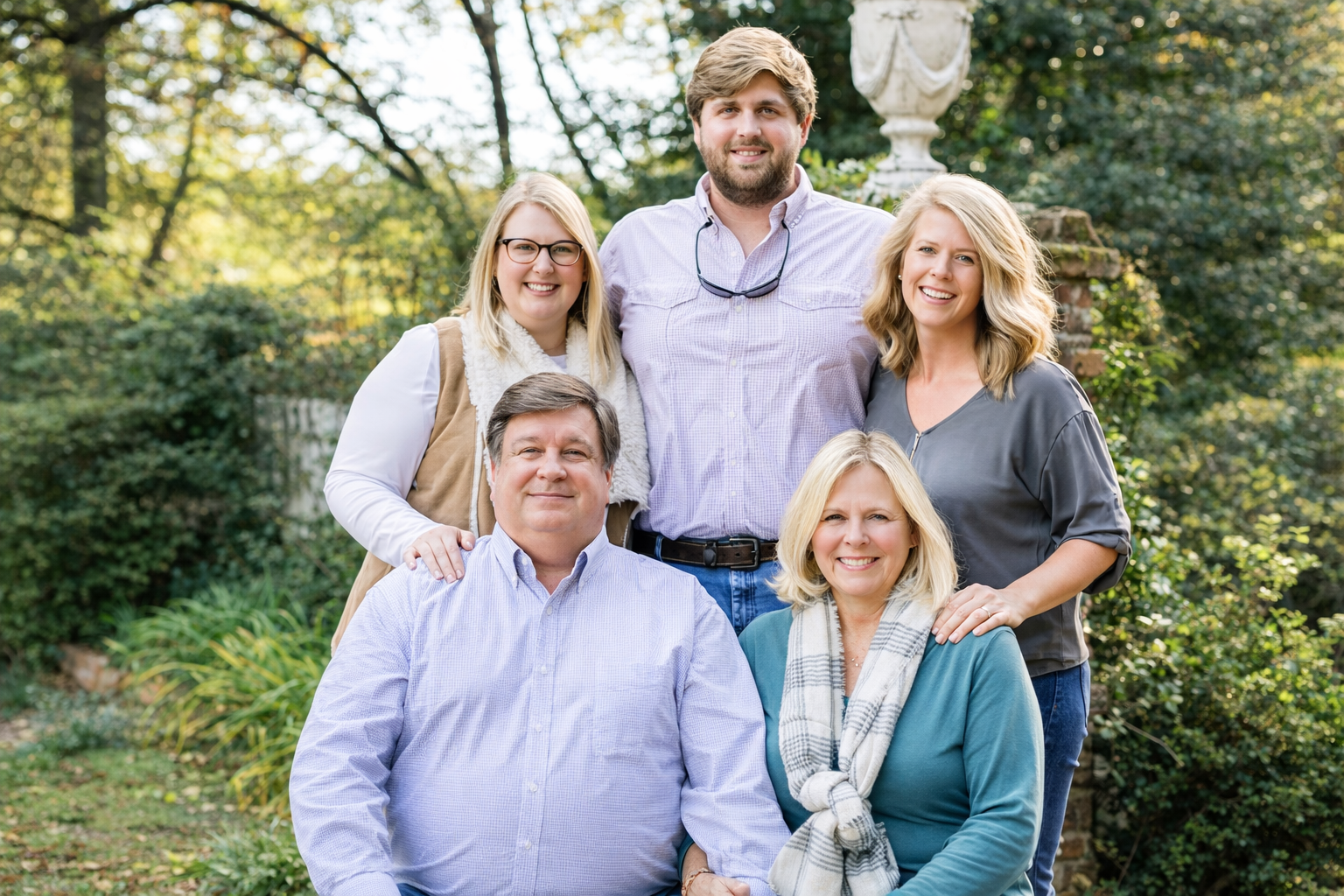 Group of six adults smiling outdoors with greenery and a stone urn in the background.