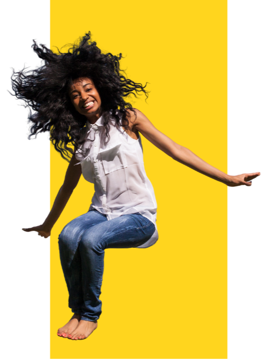A young woman with large, curly hair, smiling and sitting on an invisible surface against a yellow background, wearing a white sleeveless top and blue jeans.