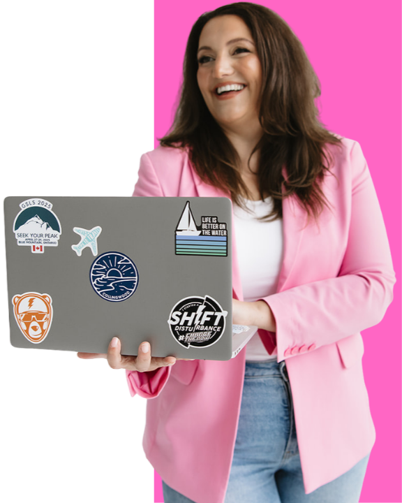 A woman with brown hair wearing a pink blazer and white shirt, holding a laptop with travel stickers, smiling against a pink background.