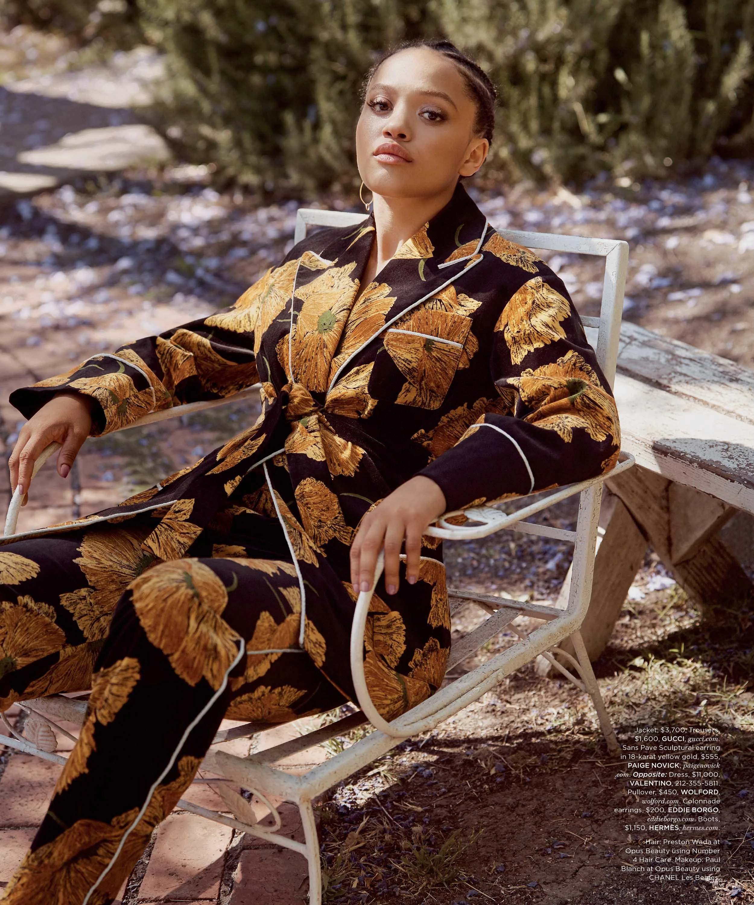 A woman sitting on a white garden chair outdoors, wearing a black outfit with a floral pattern, in front of trees and dirt ground.