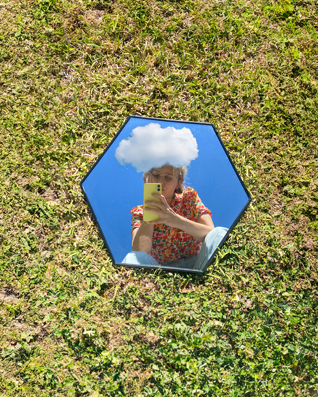 A person taking a selfie using a mirror on grass, with a cloud reflection positioned above their head, creating the illusion of having a cloud on their head. The person is wearing a colorful floral shirt and glasses, and their reflection is visible i