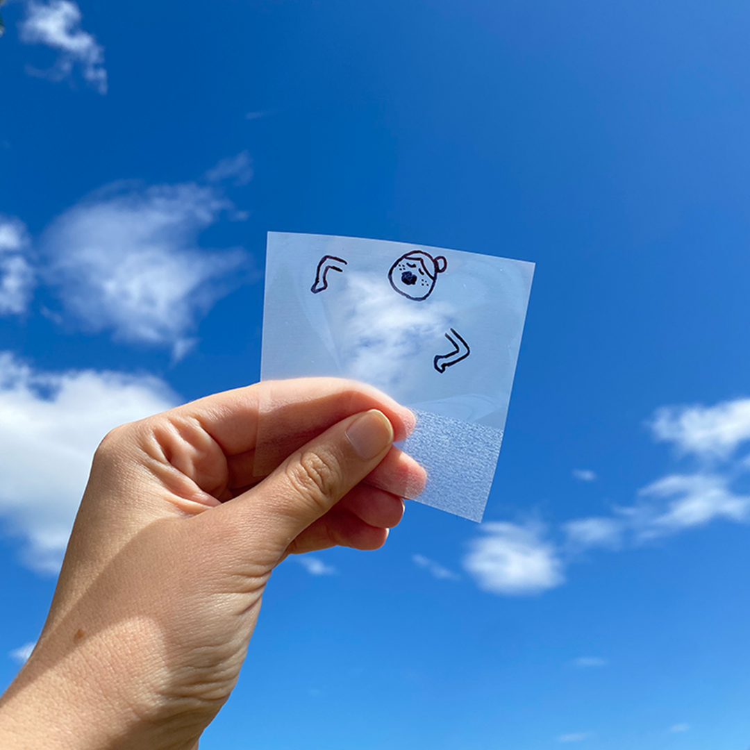 A hand holding a small transparent sheet with a simple cartoon drawing of a person wearing a hat, against a bright blue sky with some clouds.