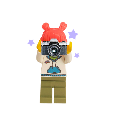 LEGO minifigure with red hair holding a camera, wearing a beige shirt with a frog graphic and olive green pants.