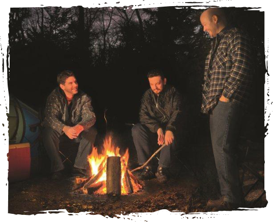 Three men sitting and standing around a campfire outdoors at night, with trees in the background.