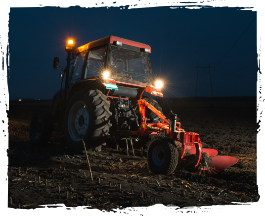 A tractor with lights on operating in a dark field at dusk or night, tilling or planting the soil.